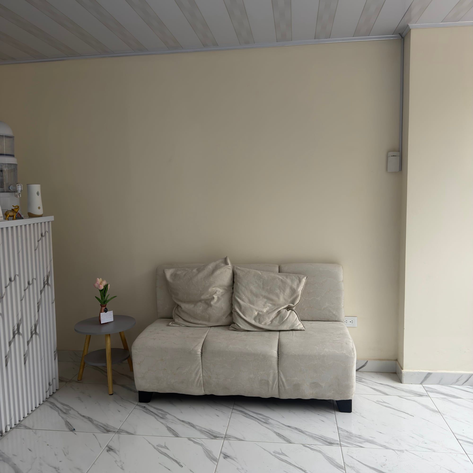 Sofá color crema con dos cojines contra la pared, al lado de una mesa auxiliar con un florero pequeño.