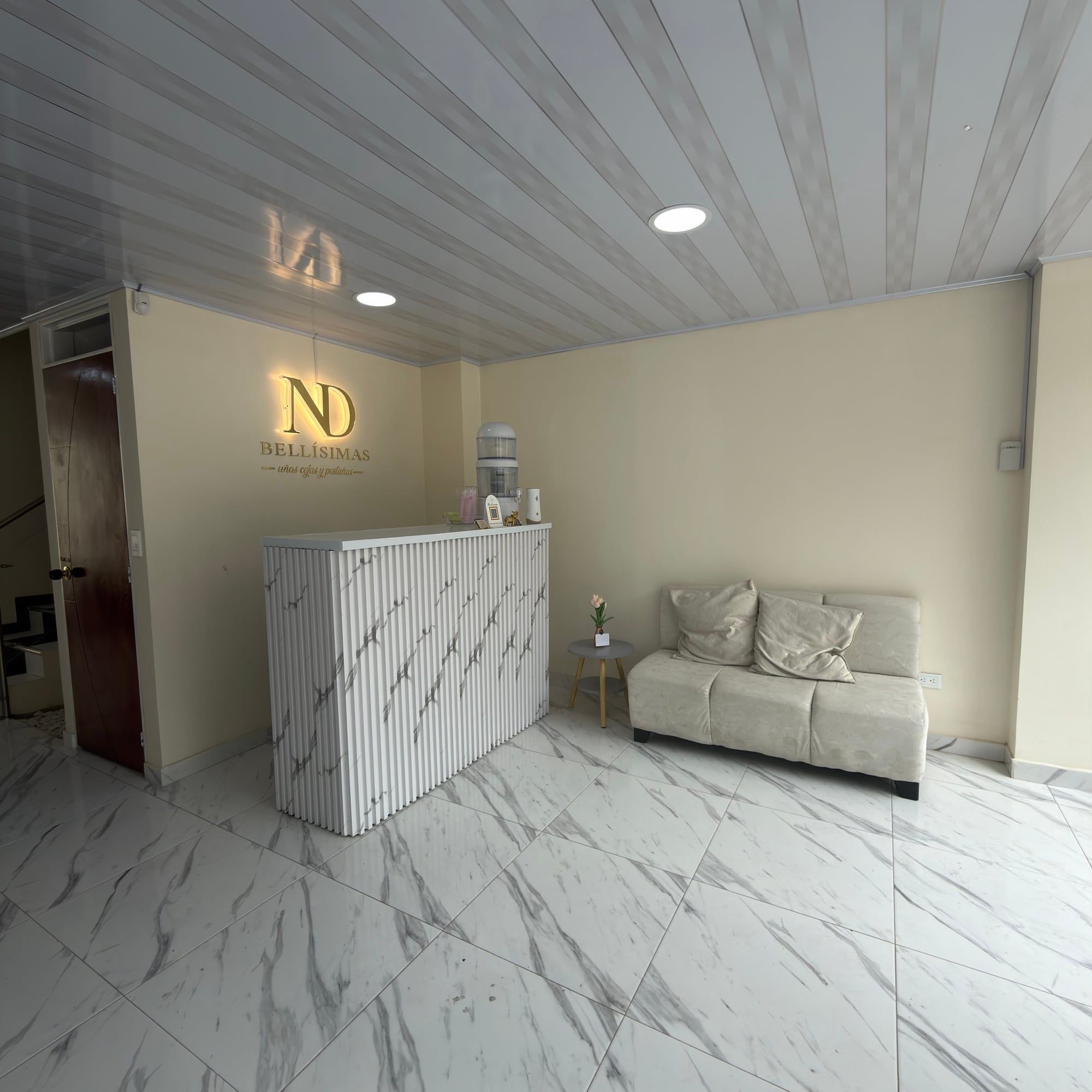 Recepción con mostrador blanco veteado y el logo ND Bellísimas iluminado en la pared; a la derecha, un sofá color crema con cojines junto a una mesa pequeña con flores.