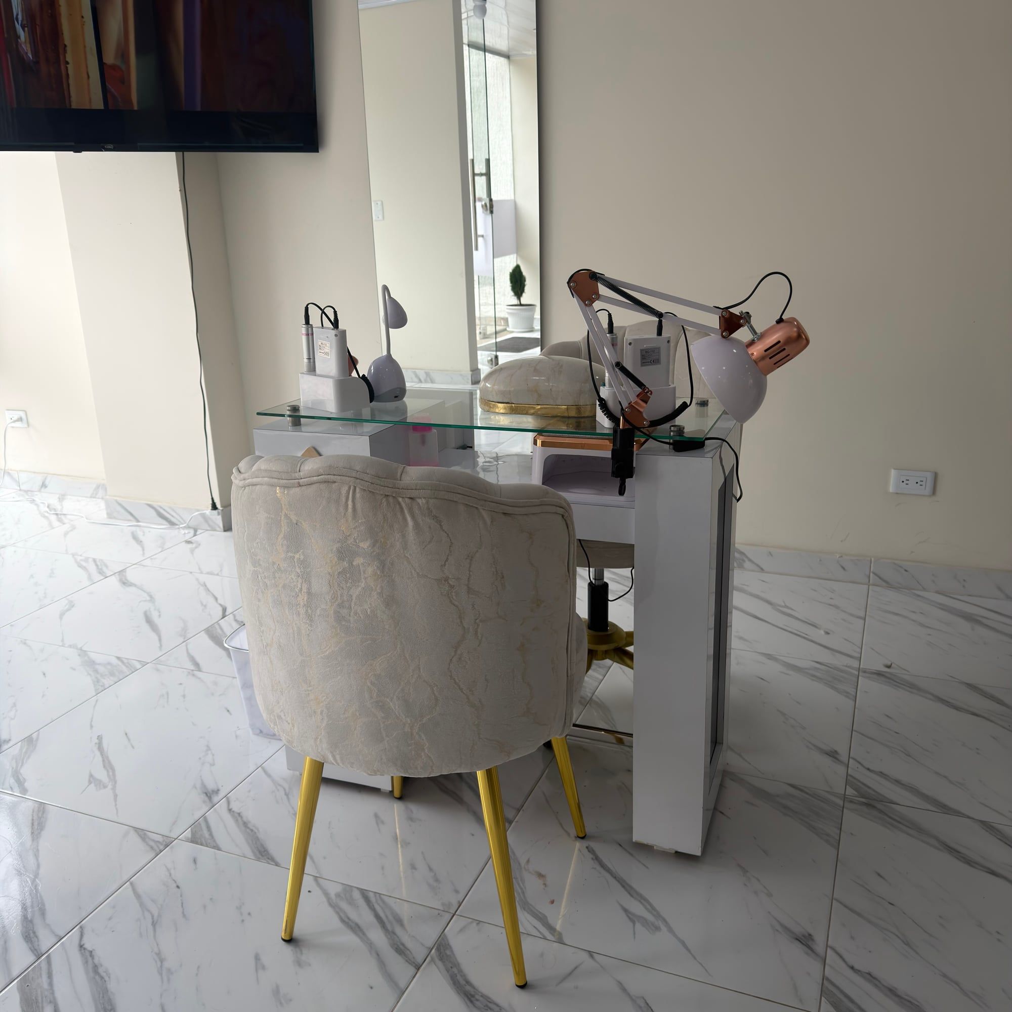 Puesto de manicure con silla tapizada color crema de patas doradas frente a una mesa blanca con lámparas, junto a un espejo de pie.