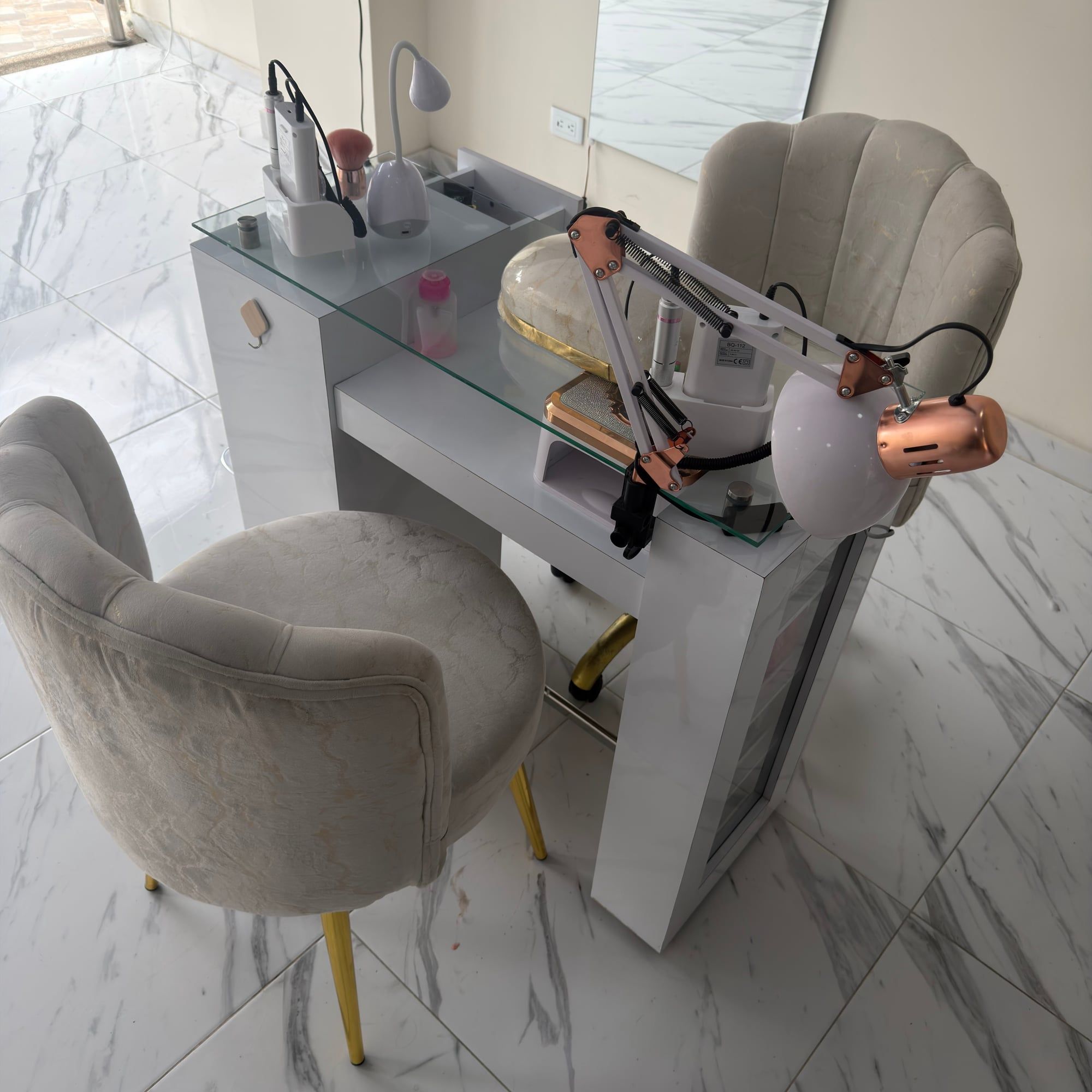 Mesa de manicure blanca con cubierta de vidrio, lámpara articulada y lámpara para uñas, frente a un sillón tapizado color crema con patas doradas.