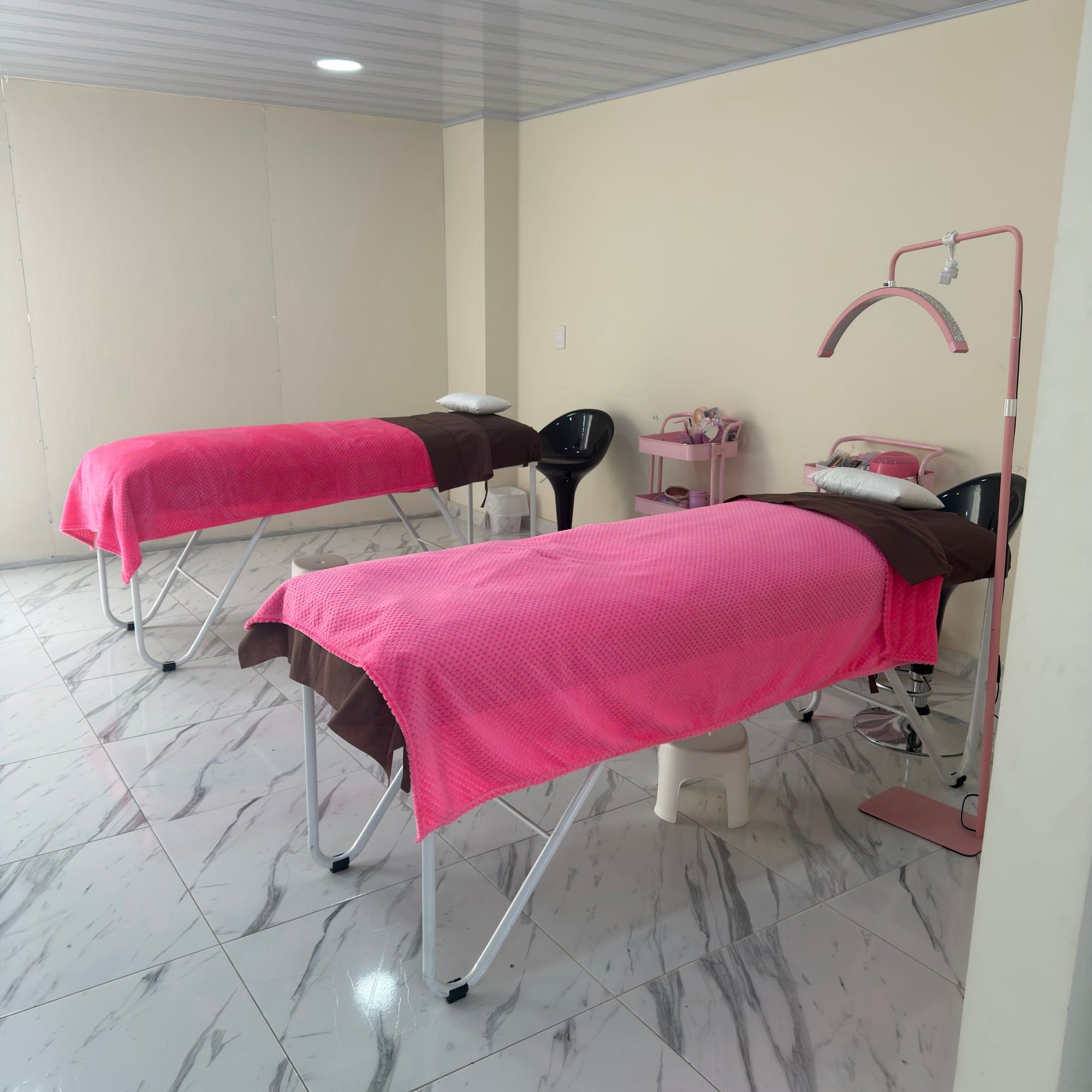 Dos camillas con cobijas rosadas y toallas cafés, una lámpara rosada en arco y carritos auxiliares rosados.