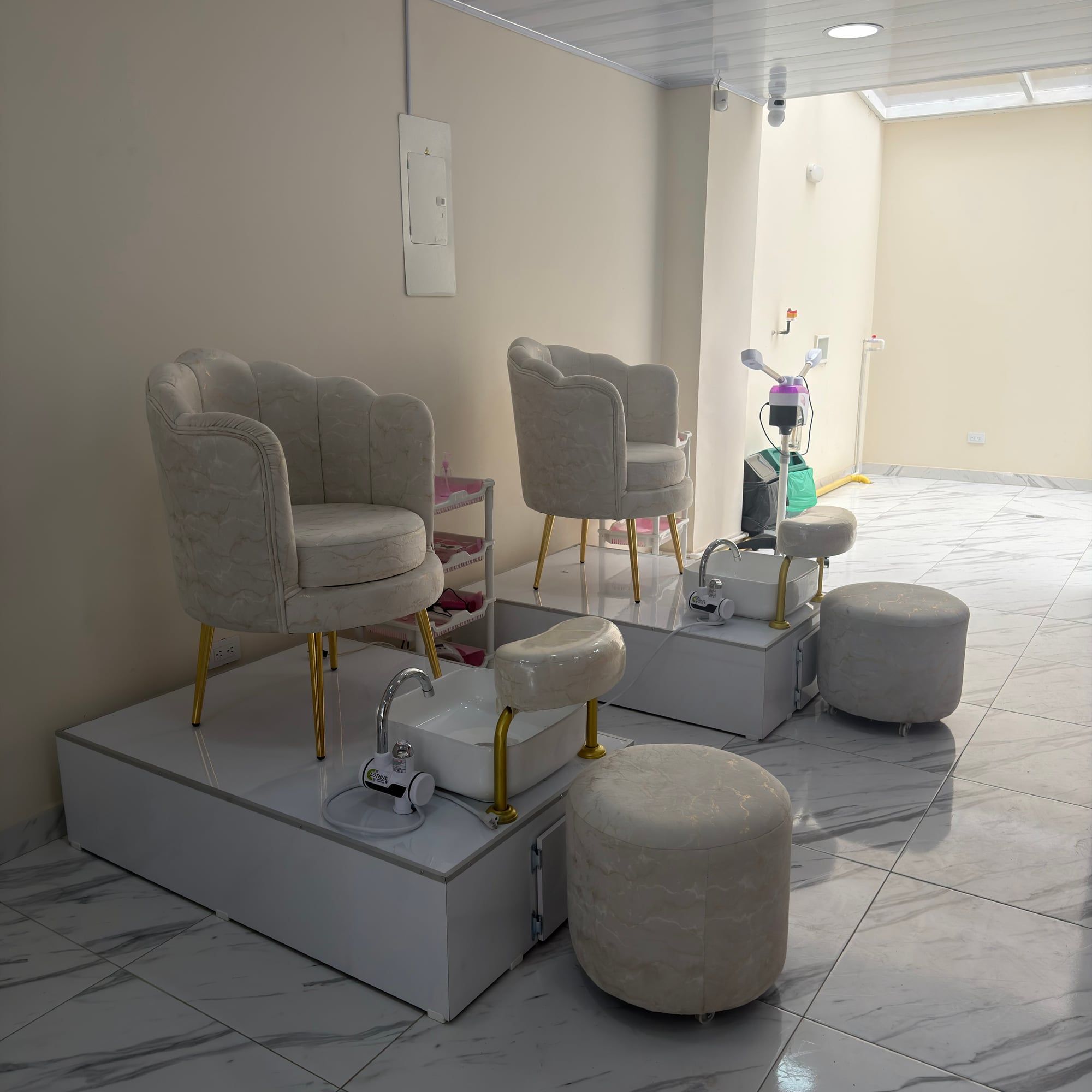 Dos sillones color crema con patas doradas sobre plataformas blancas de pedicure, con lavapiés, apoyapiés y taburetes tapizados al frente.