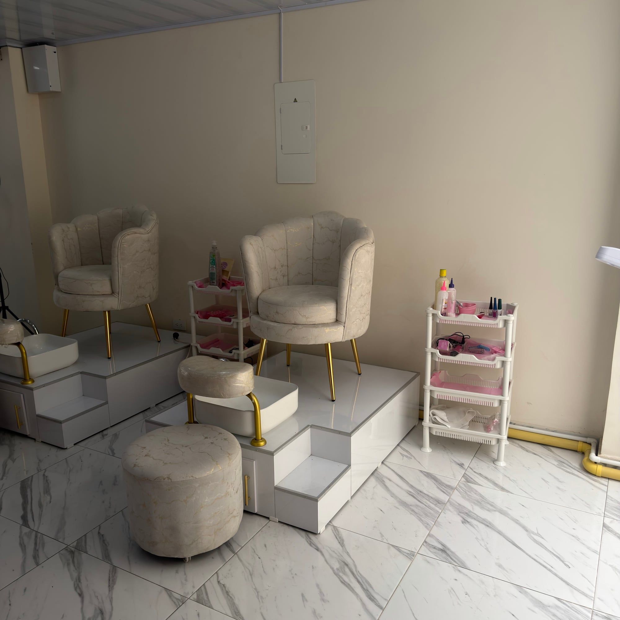 Zona de pedicure con dos sillones color crema sobre plataformas blancas con lavapiés y un organizador blanco con productos en bandejas rosadas.