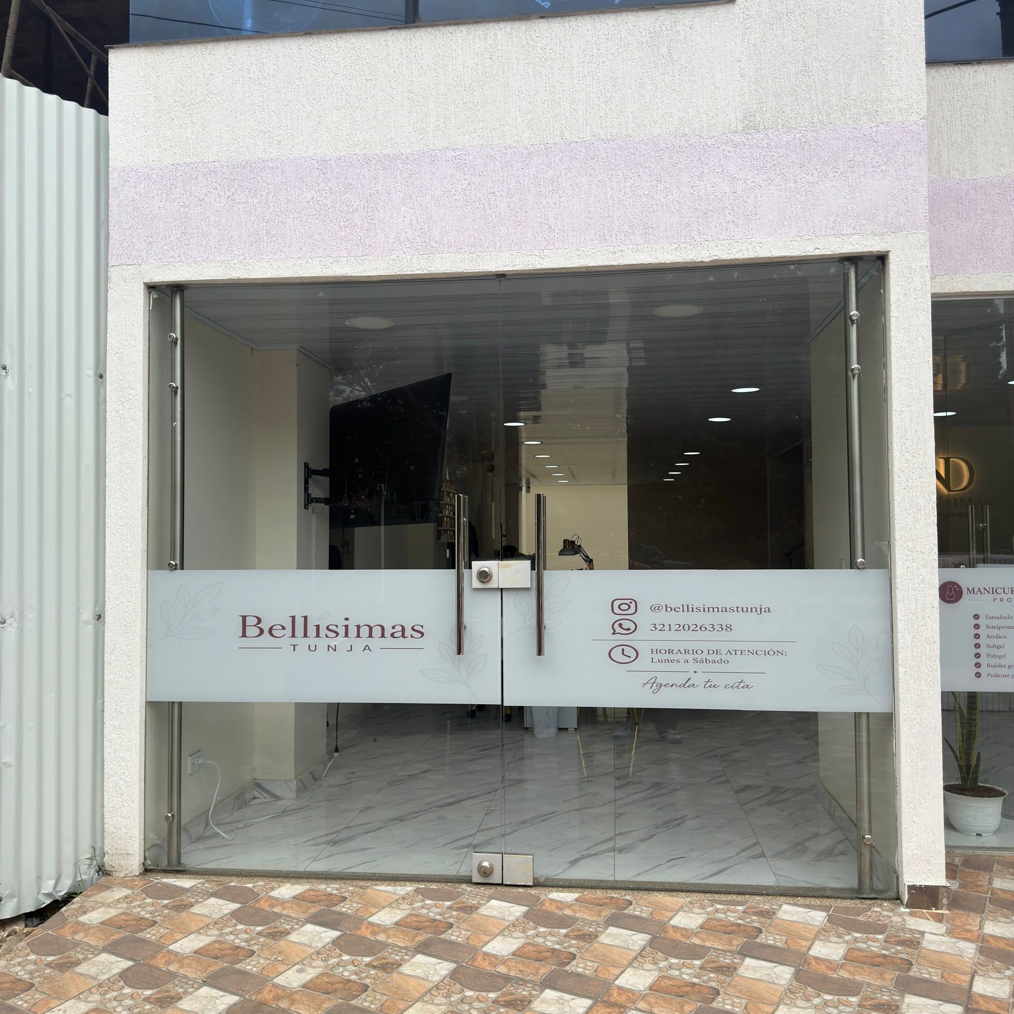 Fachada del local desde la acera: puertas de vidrio con un vinilo blanco que dice «Bellísimas Tunja», el Instagram, un número de WhatsApp y el horario de lunes a sábado.