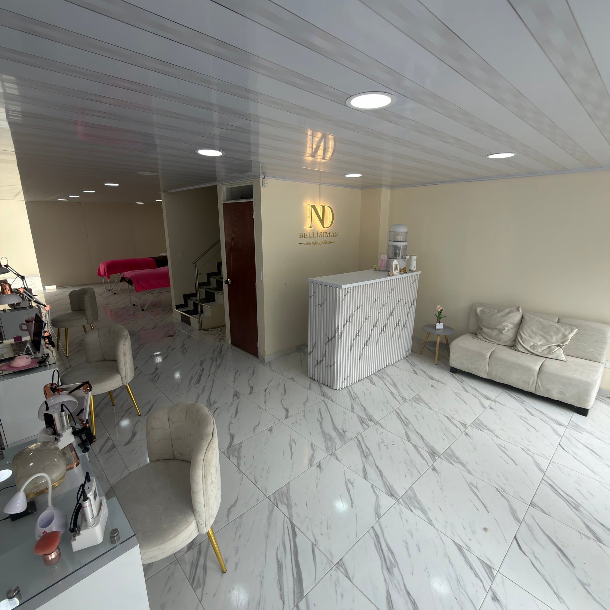 Vista amplia del interior del local: puestos de manicure con lámparas y sillas color crema de patas doradas a la izquierda, camillas con cobijas rosadas al fondo y, a la derecha, la recepción con el logo ND Bellísimas iluminado y un sofá.